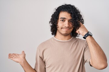 Hispanic man with curly hair standing over white background confused and annoyed with open palm showing copy space and pointing finger to forehead. think about it.