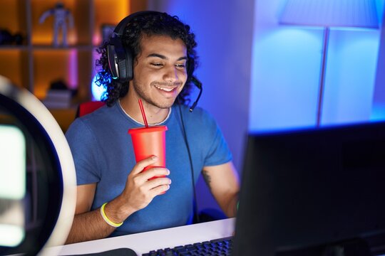 Young latin man streamer playing video game drinking beverage at gaming room