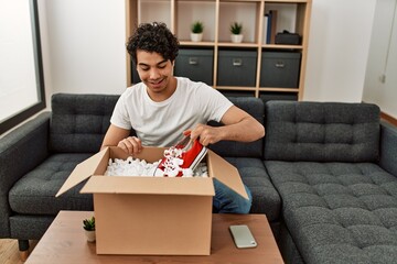 Young hispanic man unboxing sneakers of cardboard box sitting on the sofa at home.