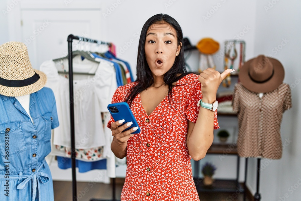 Sticker young hispanic woman working at retail boutique using smartphone surprised pointing with hand finger