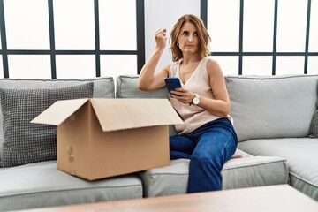 Middle age woman sitting on the sofa checking delivery package on the phone annoyed and frustrated shouting with anger, yelling crazy with anger and hand raised