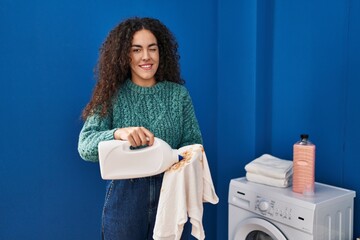 Young hispanic woman holding dirty laundry and detergent bottle winking looking at the camera with...