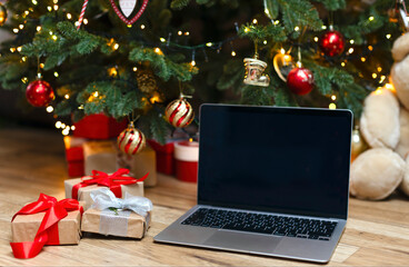 A laptop with space for a copy on a wooden background under a Christmas tree. An open laptop stands under the Christmas tree with gifts, next to them are boxes decorated with red ribbons.