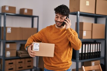 Young hispanic man ecommerce business worker talking on smartphone holding package at office