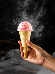 Woman's hand holding strawberry ice cream cone with smoke effect. Dark background. Conceptual...
