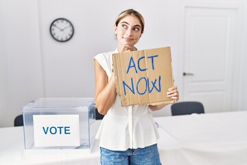 Young blonde woman at political election holding act now banner serious face thinking about question with hand on chin, thoughtful about confusing idea