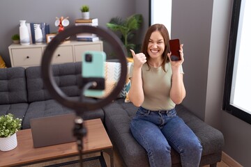 Beautiful brunette woman recording vlog tutorial with smartphone at home pointing thumb up to the...