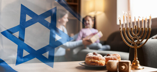 Banner with Israeli national flag and menorah, dreidels and donuts on table of happy family celebrating Hannukah at home