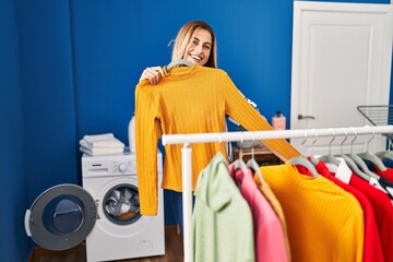 Young blonde woman smiling confident holding clothes of rack at laundry room