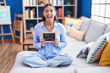 Young hispanic woman holding blackboard with new home text sitting on the sofa celebrating crazy and amazed for success with open eyes screaming excited.
