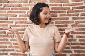 Young hispanic woman standing over bricks wall smiling showing both hands open palms, presenting and advertising comparison and balance