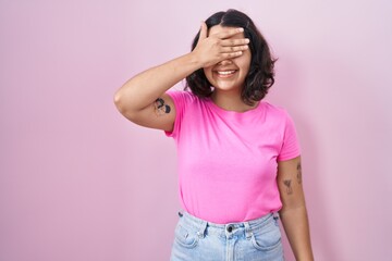Young hispanic woman standing over pink background smiling and laughing with hand on face covering eyes for surprise. blind concept.