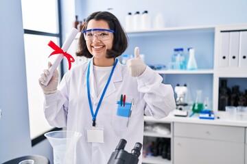 Young hispanic woman working at scientist laboratory holding degree smiling happy and positive, thumb up doing excellent and approval sign