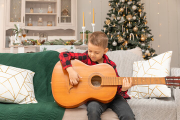 Portrait of little boy playing guitar sits on sofa against backdrop of christmas tree with garland...