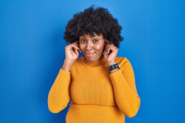 Black woman with curly hair standing over blue background covering ears with fingers with annoyed...