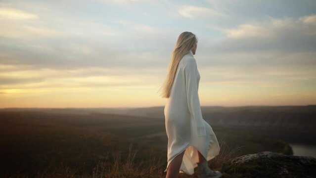 Incredibly beautiful blonde girl in a white knitted dress goes to the top of the rock above the river and raises her hands in the air to greet the sunset