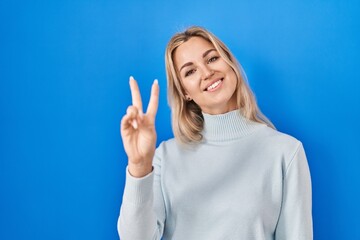 Young caucasian woman standing over blue background smiling looking to the camera showing fingers doing victory sign. number two.