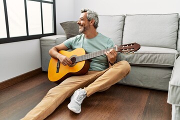 Middle age grey-haired man playing classical guitar sitting on floor at home