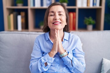 Fototapeta na wymiar Middle age hispanic woman sitting on the sofa at home praying with hands together asking for forgiveness smiling confident.