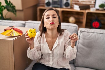 Middle age hispanic woman holding piggy bank at new home looking at the camera blowing a kiss being lovely and sexy. love expression.