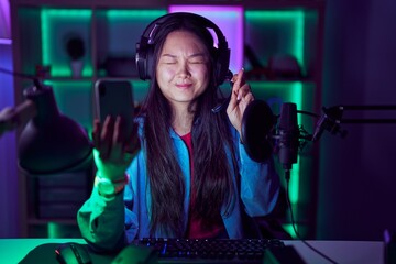Young asian woman playing video games with smartphone gesturing finger crossed smiling with hope and eyes closed. luck and superstitious concept.