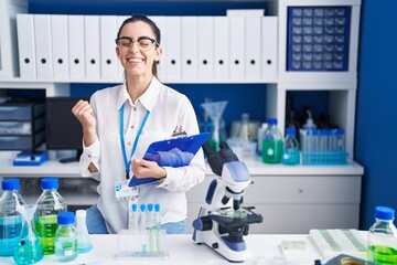 Young brunette woman working at scientist laboratory very happy and excited doing winner gesture with arms raised, smiling and screaming for success. celebration concept.