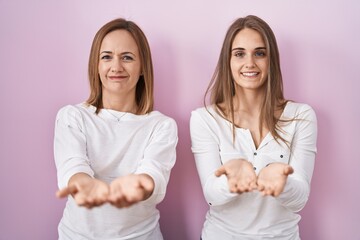 Middle age mother and young daughter standing over pink background smiling with hands palms together receiving or giving gesture. hold and protection