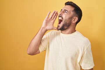 Handsome hispanic man standing over yellow background shouting and screaming loud to side with hand on mouth. communication concept.