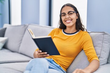 Young african american woman reading book sitting on sofa at home