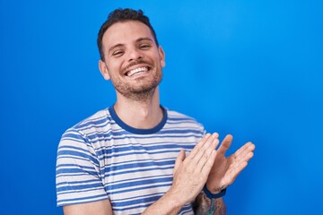Young hispanic man standing over blue background clapping and applauding happy and joyful, smiling...