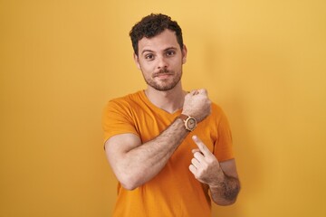 Young hispanic man standing over yellow background in hurry pointing to watch time, impatience, looking at the camera with relaxed expression