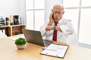 Senior man working at the office using computer laptop looking at the watch time worried, afraid of...