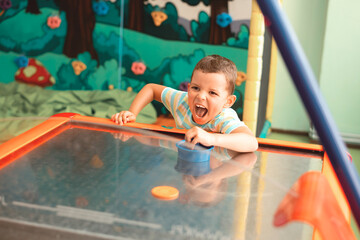 Cheerful little boy plays air hockey and really wants to win. Makes different grimaces