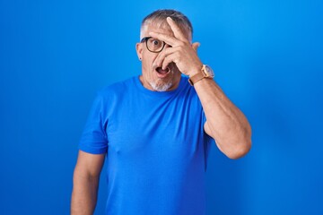 Hispanic man with grey hair standing over blue background peeking in shock covering face and eyes with hand, looking through fingers with embarrassed expression.