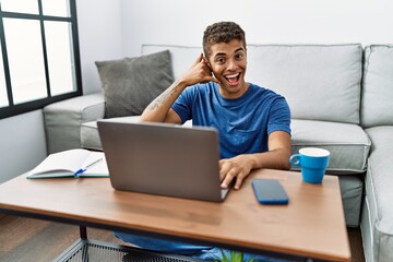 Young handsome hispanic man using laptop sitting on the floor smiling doing phone gesture with hand and fingers like talking on the telephone. communicating concepts.