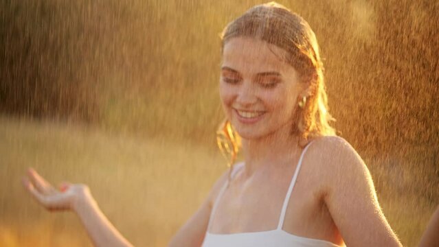 Happy blonde woman having fun in sunny park smiling, fooling around, laughing. Slow Motion. Beautiful caucasian girl dancing under the summer rain. wet hair and clothes. enjoying with drops