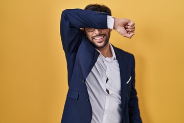 Handsome latin man standing over yellow background covering eyes with arm smiling cheerful and funny. blind concept.