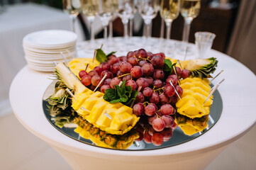 A variety of fruits, grapes, pineapple lie on a mirrored tray on a table in a restaurant. Food photography, banquet, buffet.