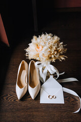 Wedding accessories, details, bride's set: beige shoes, a bottle of perfume, an envelope made of paper with sealing wax, an invitation with gold rings, a bouquet of reeds, wild flowers.