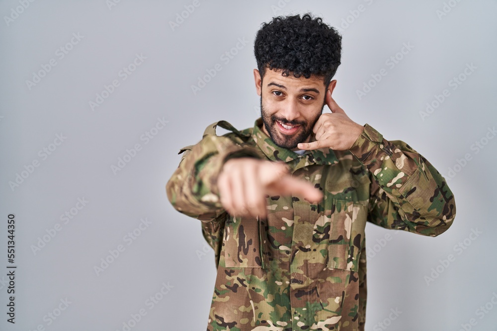 Poster arab man wearing camouflage army uniform smiling doing talking on the telephone gesture and pointing