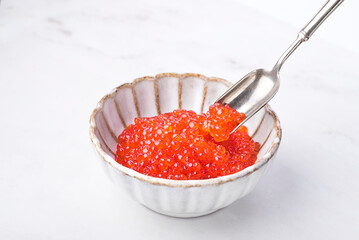 Red caviar in a beautiful ceramic bowl in the form of a flower with a silver spoon on a white  marble background. Seafood delicacies