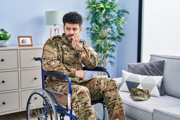 Arab man wearing camouflage army uniform sitting on wheelchair looking stressed and nervous with hands on mouth biting nails. anxiety problem.