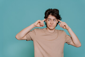 Pensive caucasian teen touches his temples by index fingers looks aside bites lip standing against blue studio backdrop with copy space. Thoughtful Italian boy in beige t-shirt isolated. Mockup. .