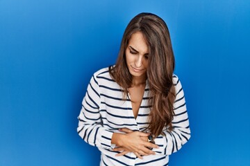 Young hispanic woman standing over blue isolated background with hand on stomach because indigestion, painful illness feeling unwell. ache concept.