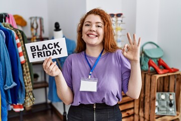 Young redhead woman holding black friday banner at retail shop doing ok sign with fingers, smiling friendly gesturing excellent symbol
