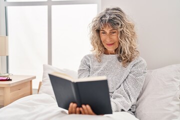 Middle age woman reading book sitting on bed at bedroom