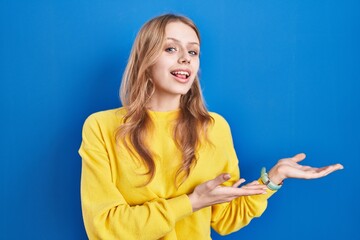 Young caucasian woman standing over blue background pointing aside with hands open palms showing copy space, presenting advertisement smiling excited happy