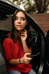 Fototapeta na wymiar Attractive pretty european woman with green eyes wearing red shirt using smartphone while sitting in the car. Outdoor portrait of successful woman