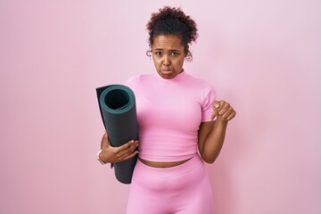 Young hispanic woman with curly hair holding yoga mat over pink background pointing down looking sad and upset, indicating direction with fingers, unhappy and depressed.