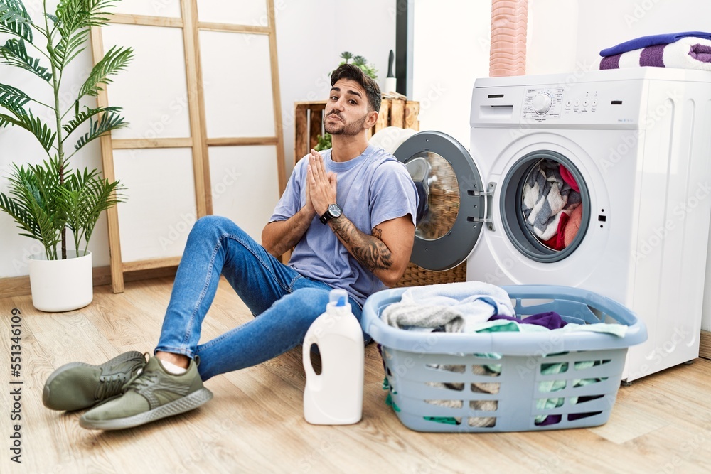 Wall mural Young hispanic man putting dirty laundry into washing machine praying with hands together asking for forgiveness smiling confident.